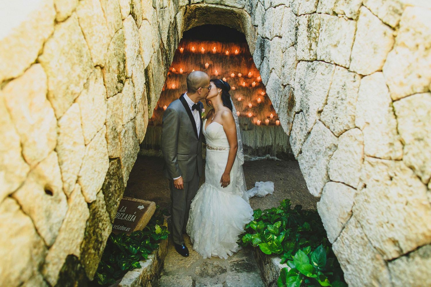 Bride and groom share a romantic kiss in a stone archway lit by warm glowing lanterns at their wedding.