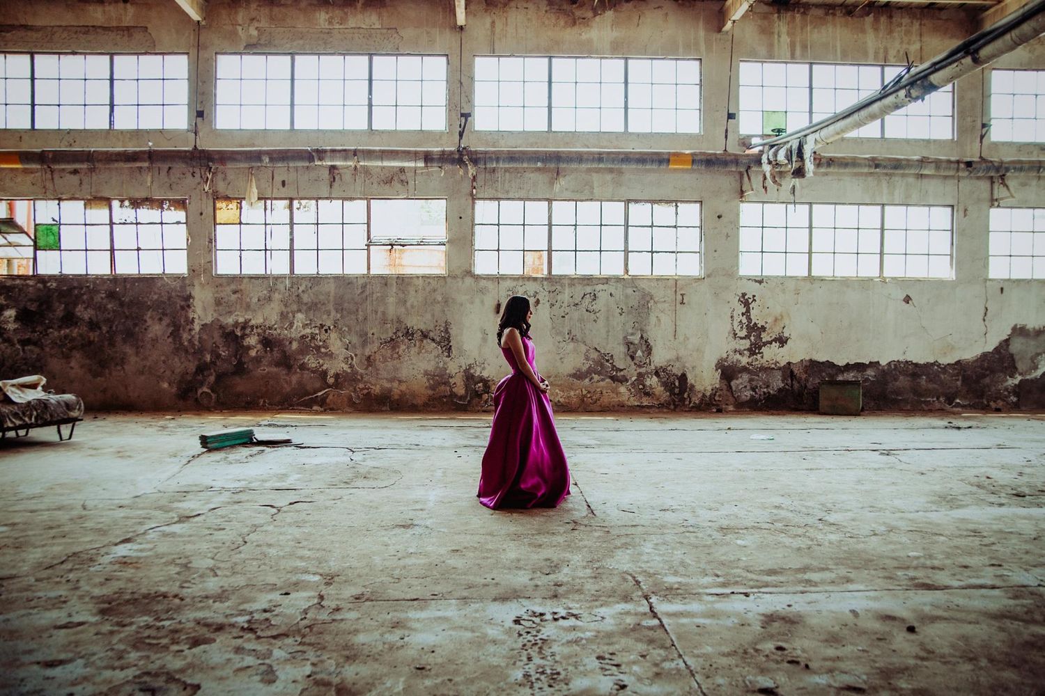 Woman in elegant magenta gown standing in abandoned industrial warehouse with large windows and weathered walls.