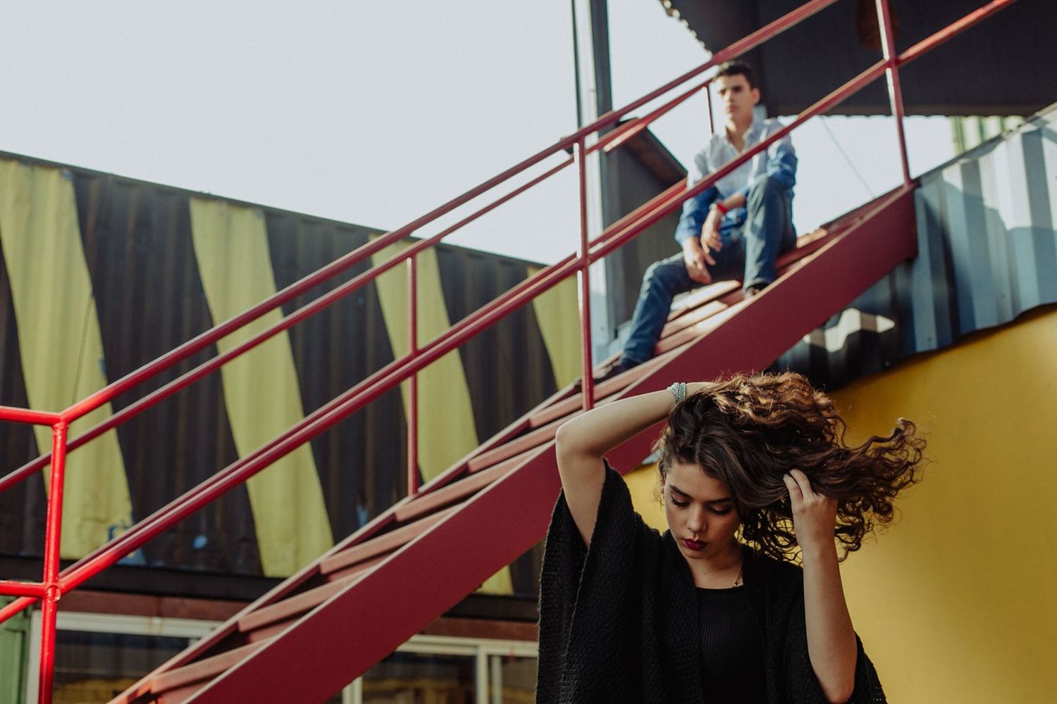 Young woman with curly hair poses near red metal stairs while a man sits above her on an urban rooftop.