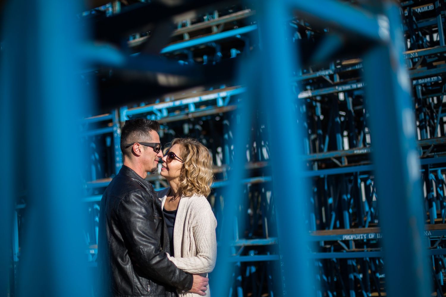 Couple embracing romantically outdoors, framed by blue metal structures, man in leather jacket and woman in beige coat.