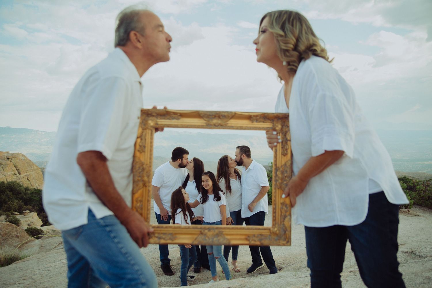 Family holding ornate gold frame outdoors in desert, reflecting group portrait in white clothing.