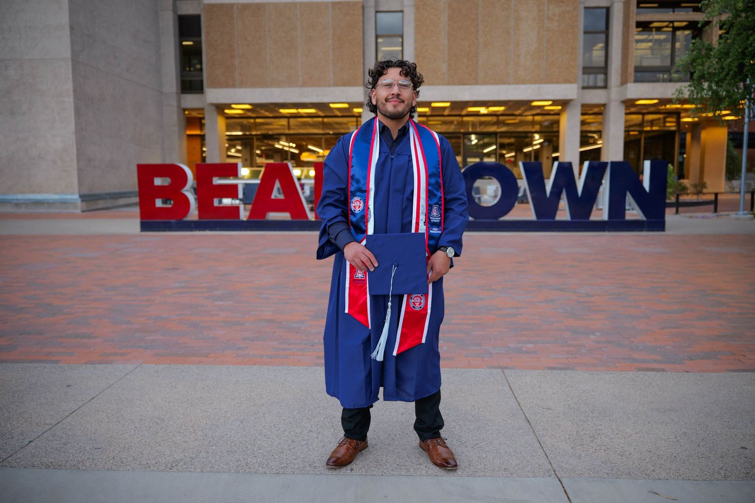 Graduate in blue robe stands before Beantown sign at Boston university graduation ceremony.