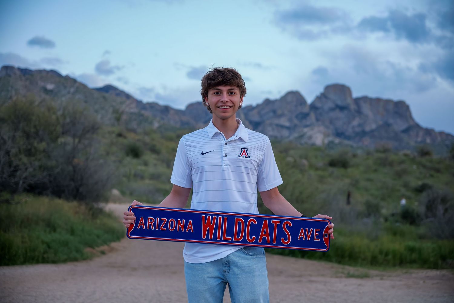 Young man holding an Arizona Wildcats banner outdoors with rocky mountain landscape behind him.
