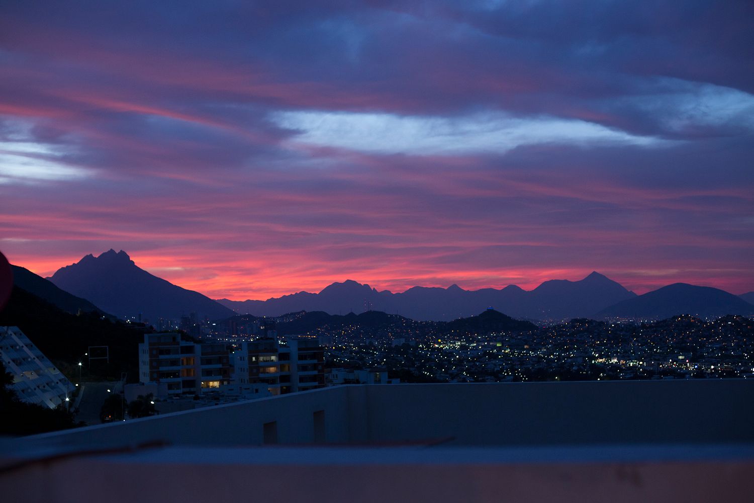 Stunning pink and purple sunset over mountain skyline with city lights glowing below dramatic cloudy sky.