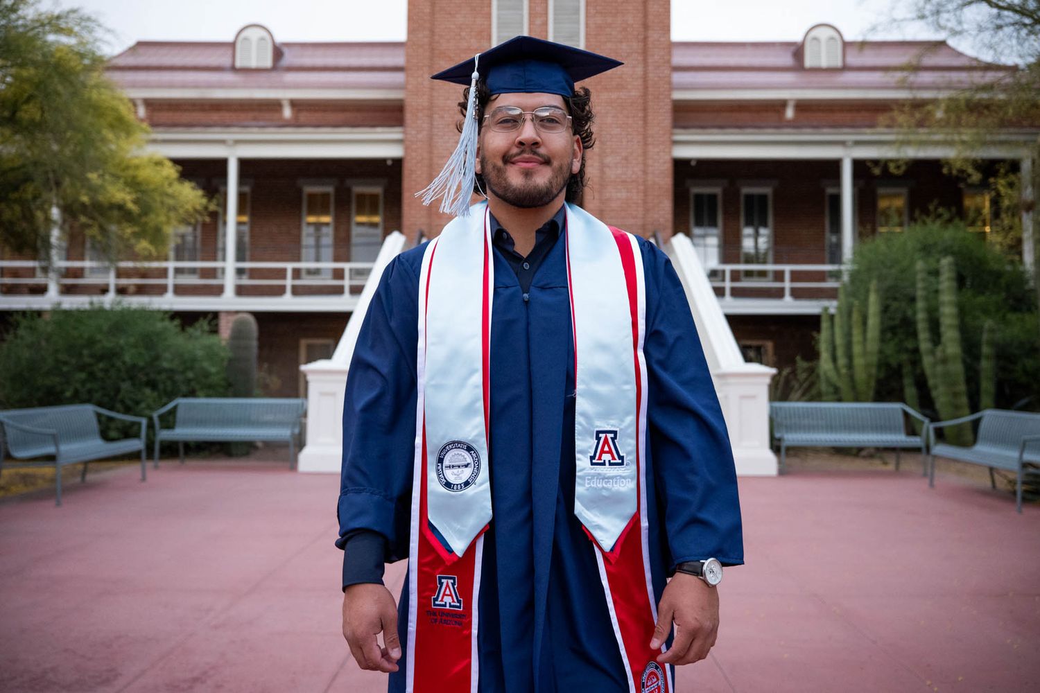 University of Arizona graduate in blue cap and gown with red and white stole stands proudly on campus.