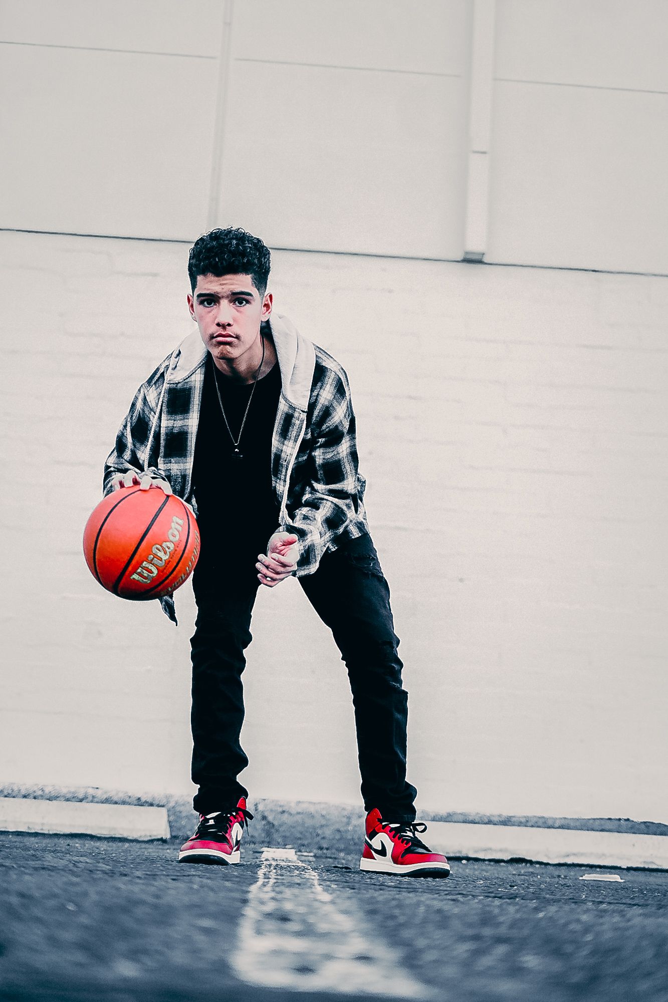 Teen boy dribbling Wilson basketball outdoors wearing plaid shirt, black jeans, and red Nike Air Jordans.