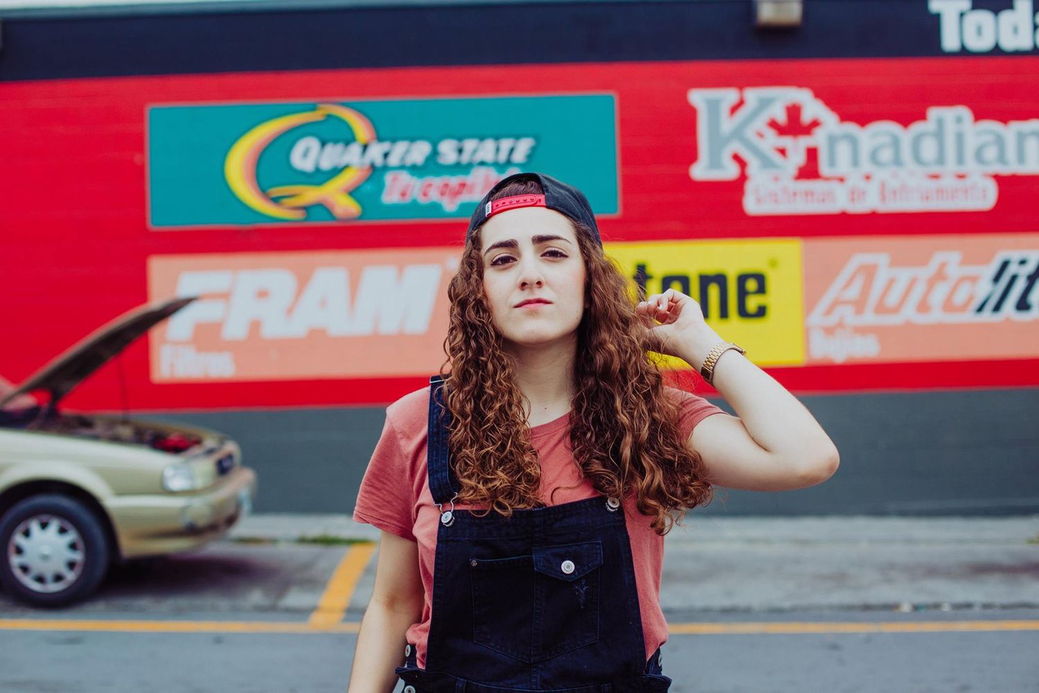 Young woman with curly hair in overalls and red cap poses in front of colorful auto shop signs.