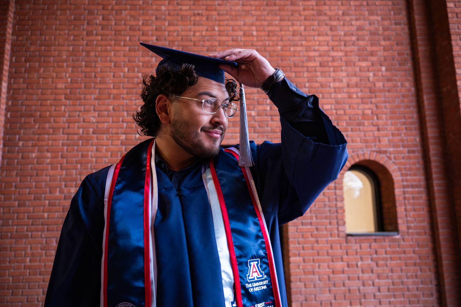 University of Arizona graduate in blue cap and gown smiles while adjusting mortarboard in front of brick building.