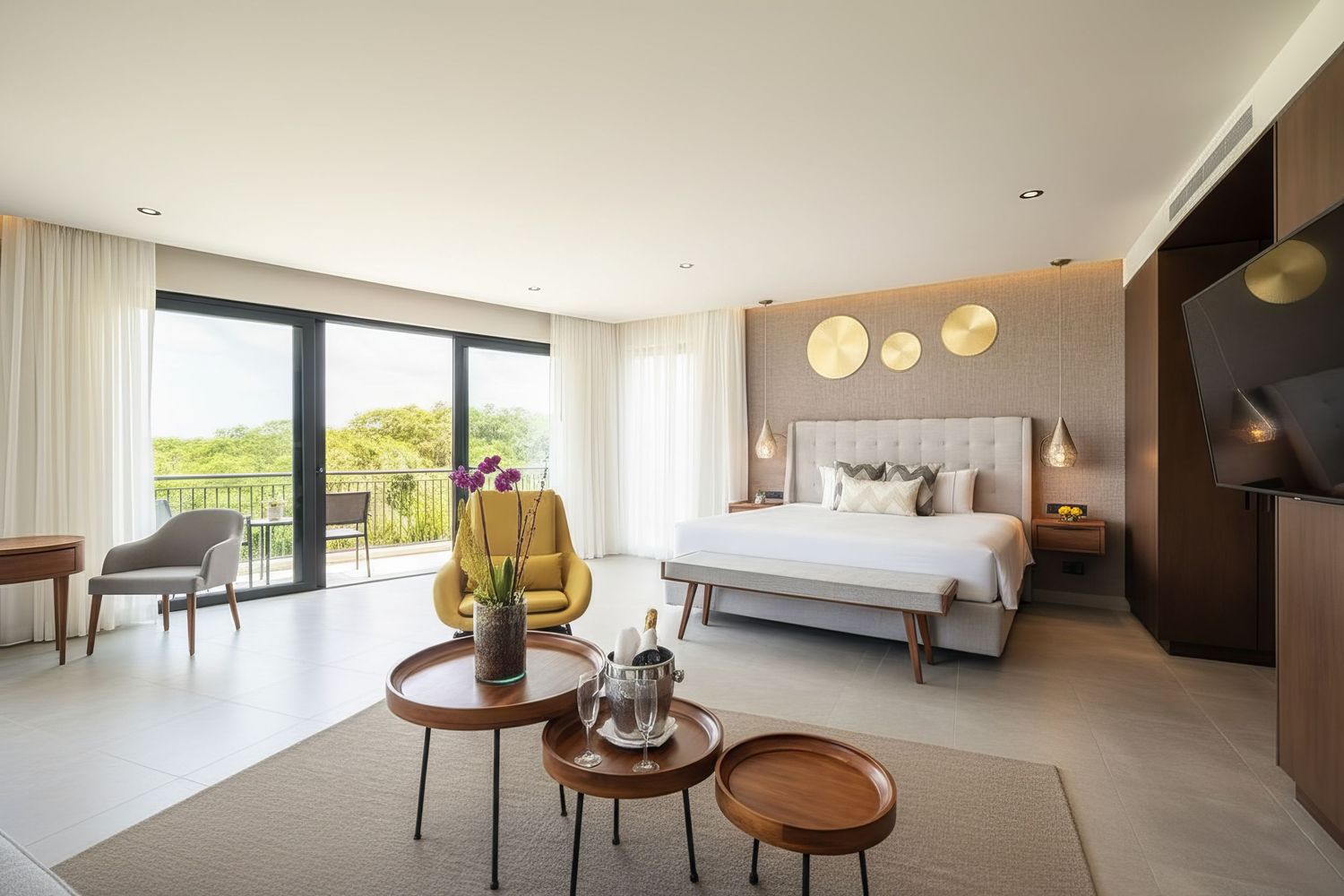 Elegant hotel suite with a king bed, yellow accent chair, round wooden tables, and a balcony with garden views.