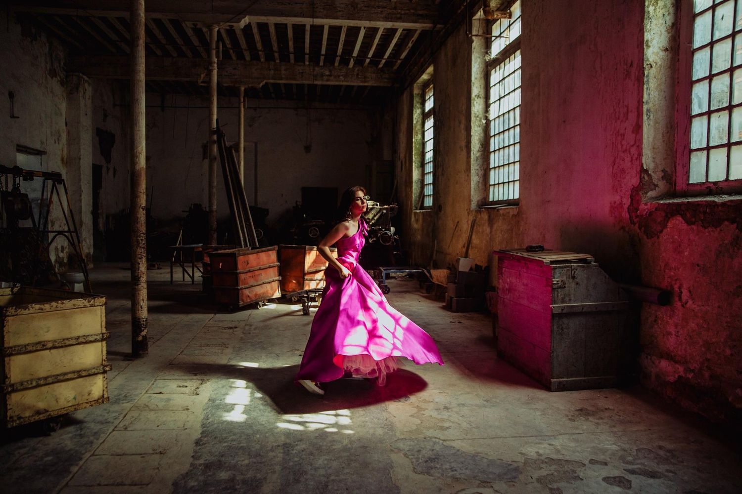 Woman in vibrant pink gown dancing gracefully in a rustic, abandoned industrial space with colorful lighting.