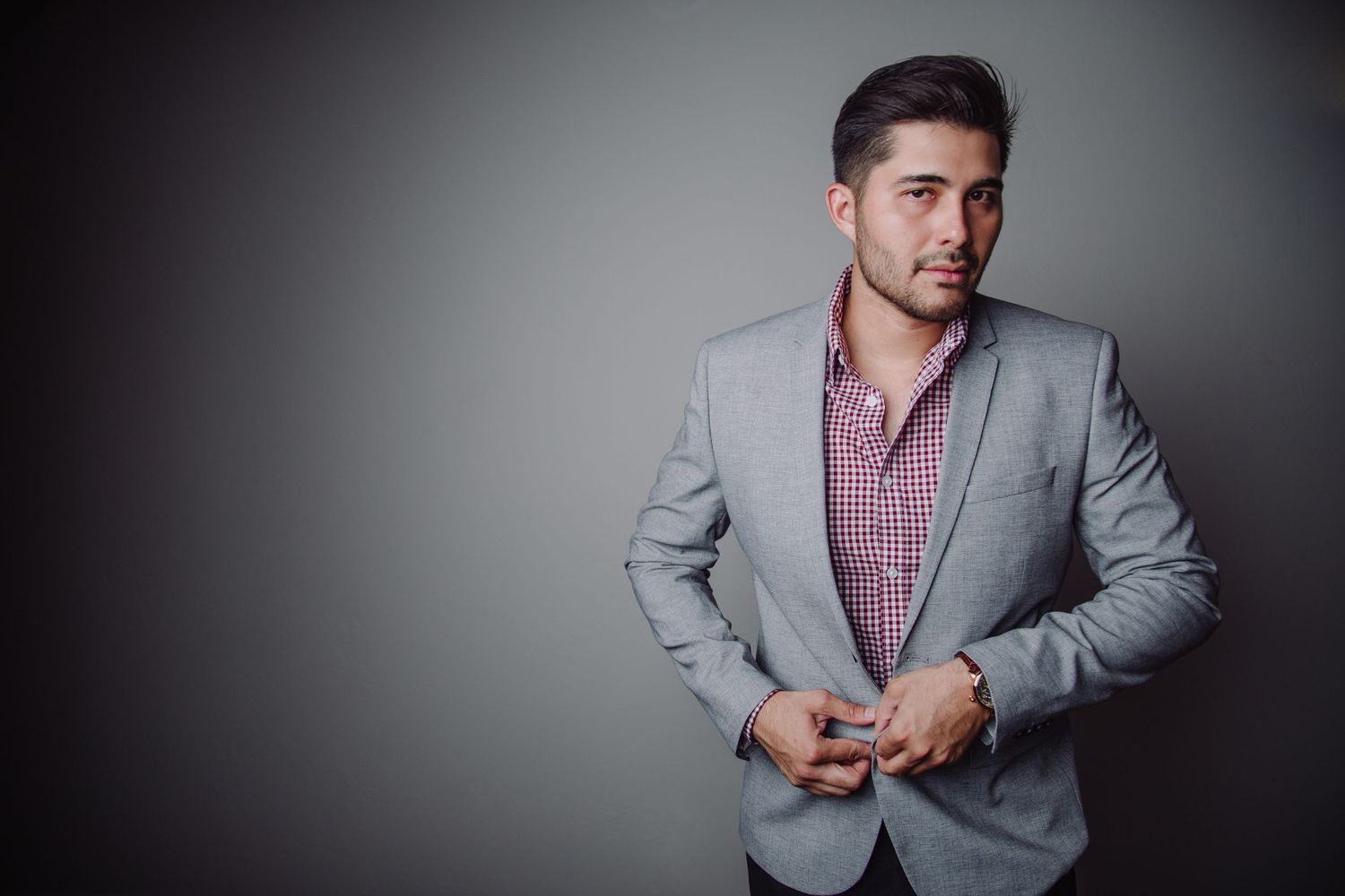 Man in gray blazer and plaid shirt standing confidently against gray background.