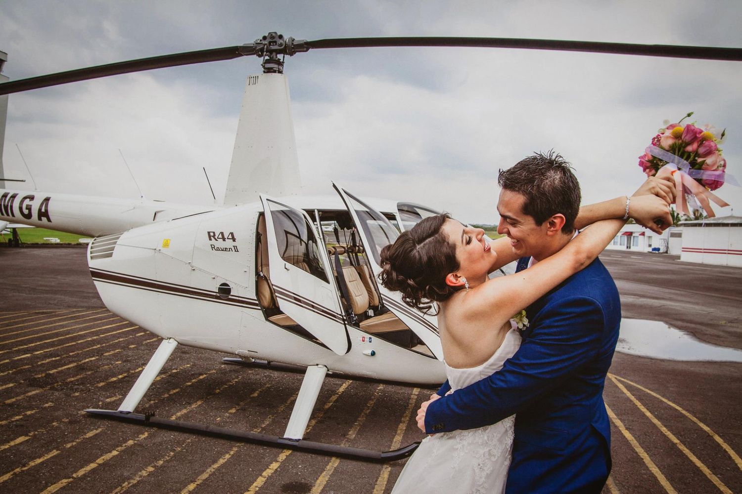Newlyweds embrace and kiss beside a white Robinson R44 helicopter on their wedding day.