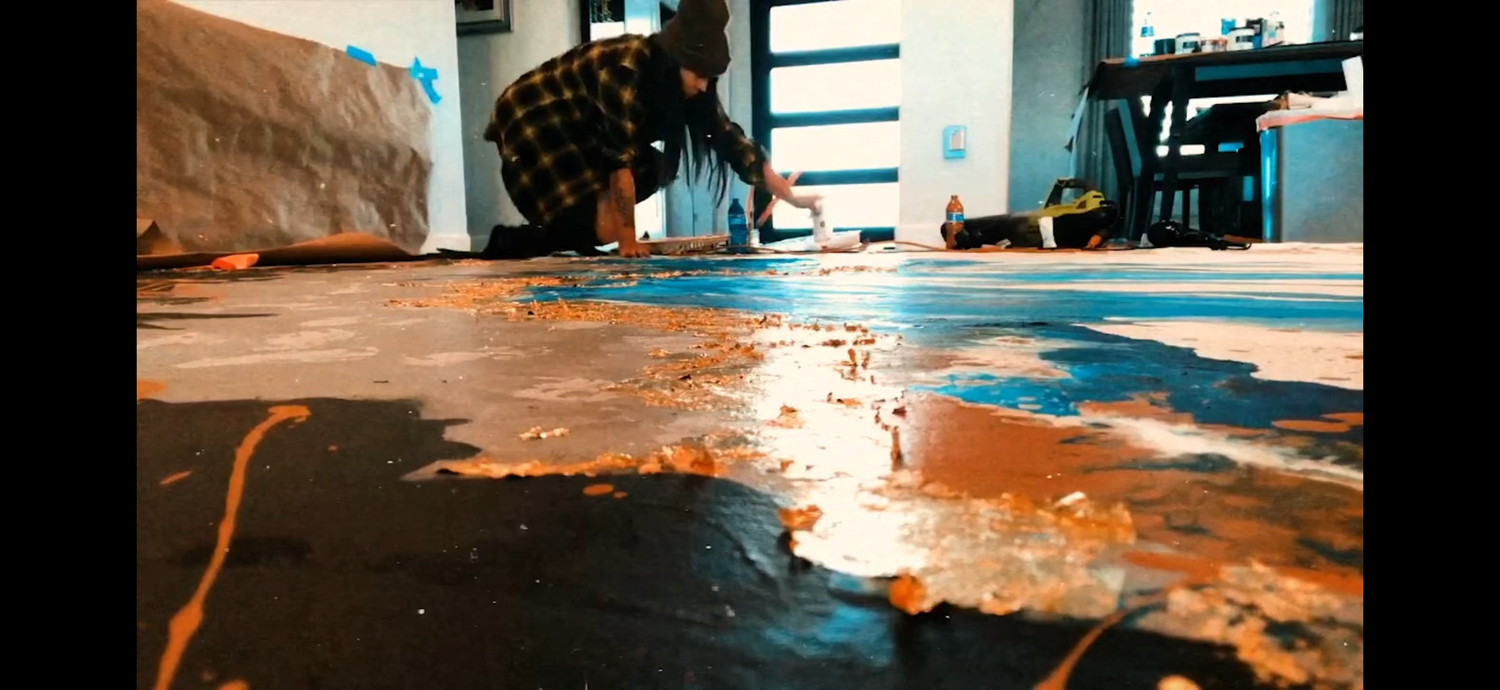 Artist working on a large abstract painting on the floor with vibrant blue and orange paint splashes.