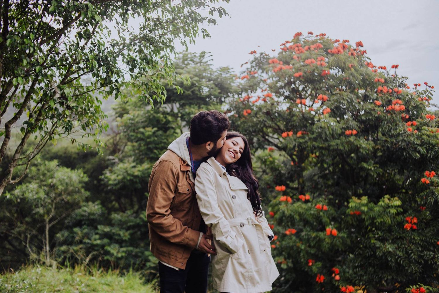 A romantic couple embraces outdoors surrounded by lush green trees and vibrant red flowers in a serene garden setting.