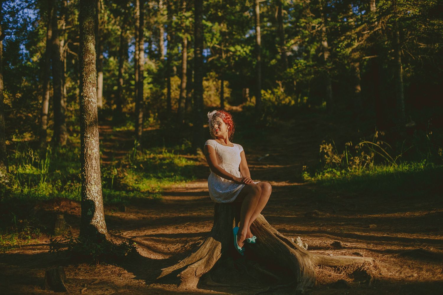 A young woman in a white dress sits peacefully on a log in a sunlit forest.