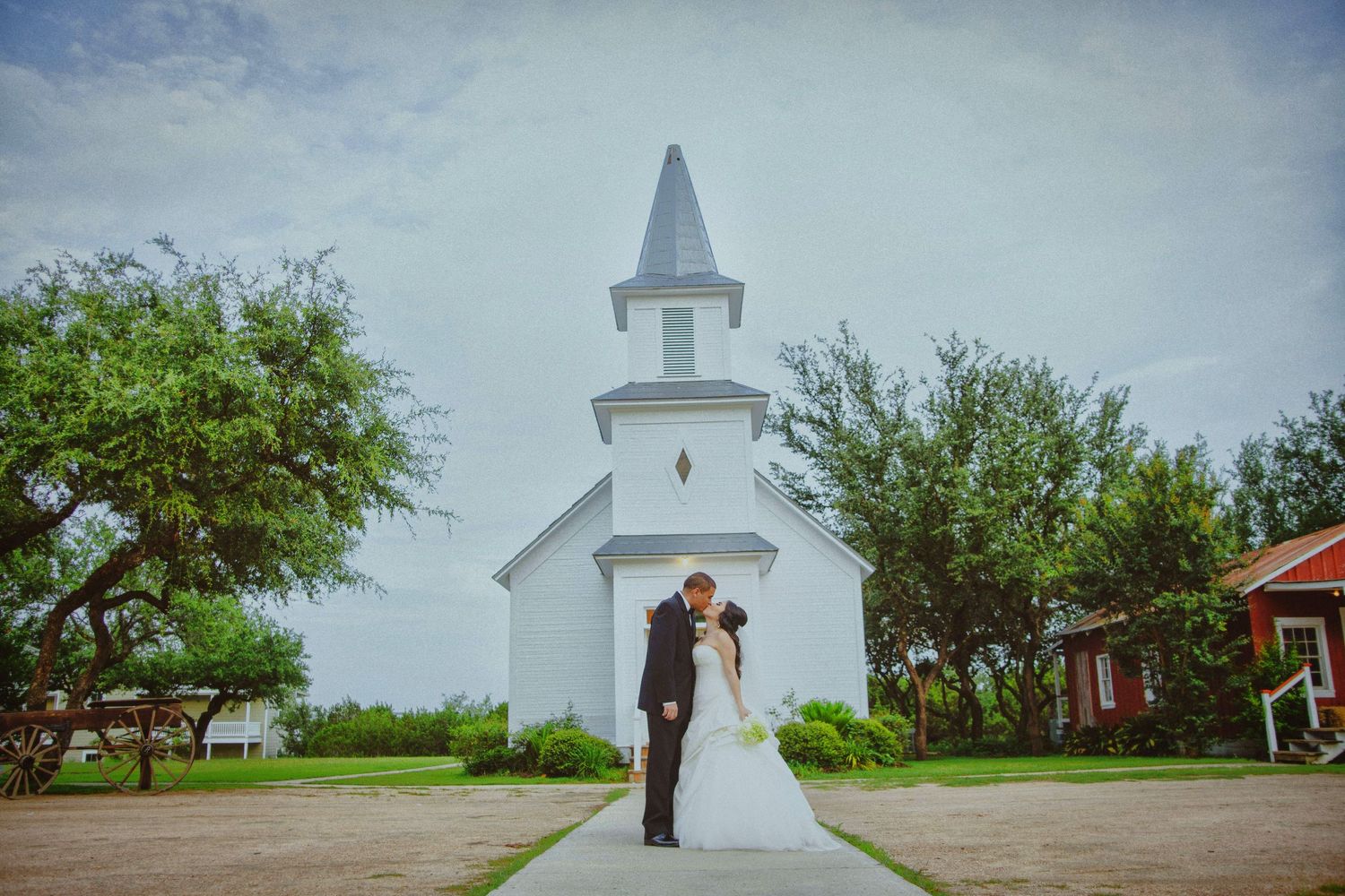 Wedding couple kisses on pathway before charming white chapel with steeple surrounded by trees.