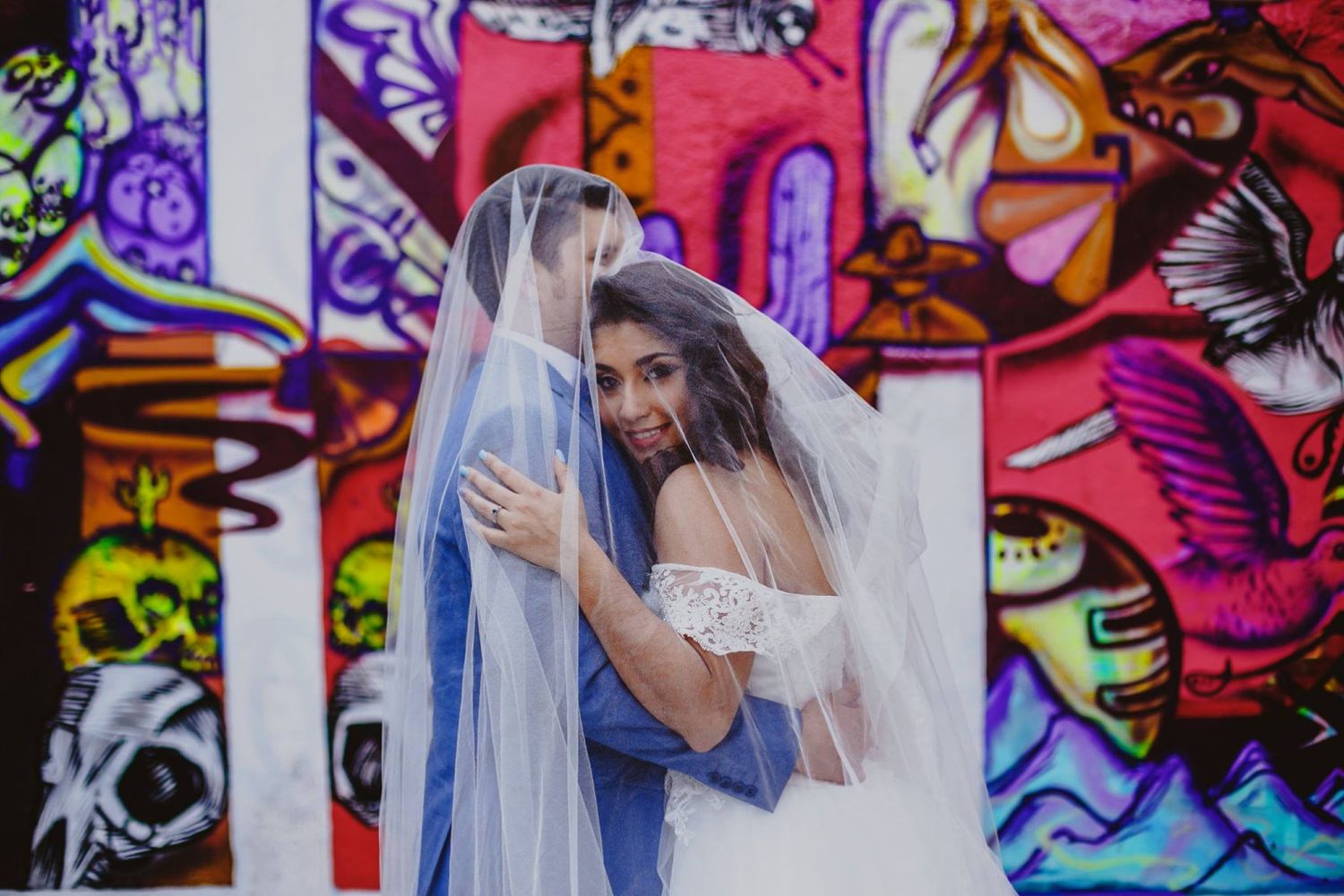 Bride and groom share a romantic moment under a wedding veil in front of a vibrant colorful graffiti wall.