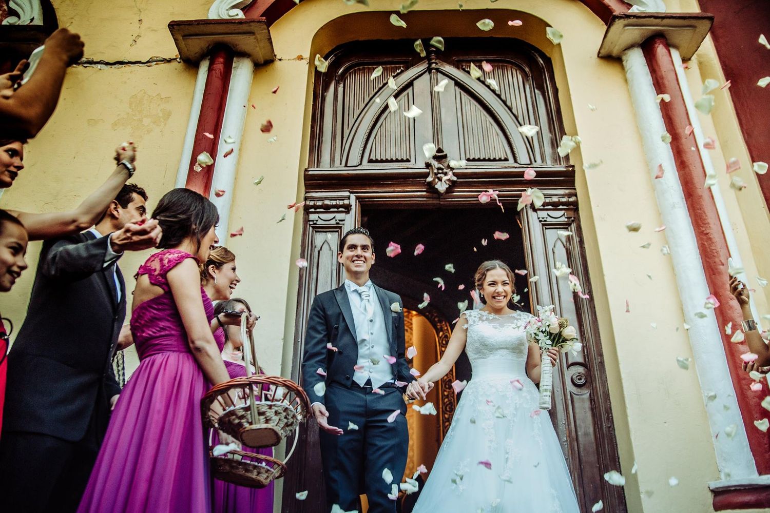 Joyful newlyweds exit a church as guests toss flower petals in celebration, with bridesmaids in pink dresses nearby.