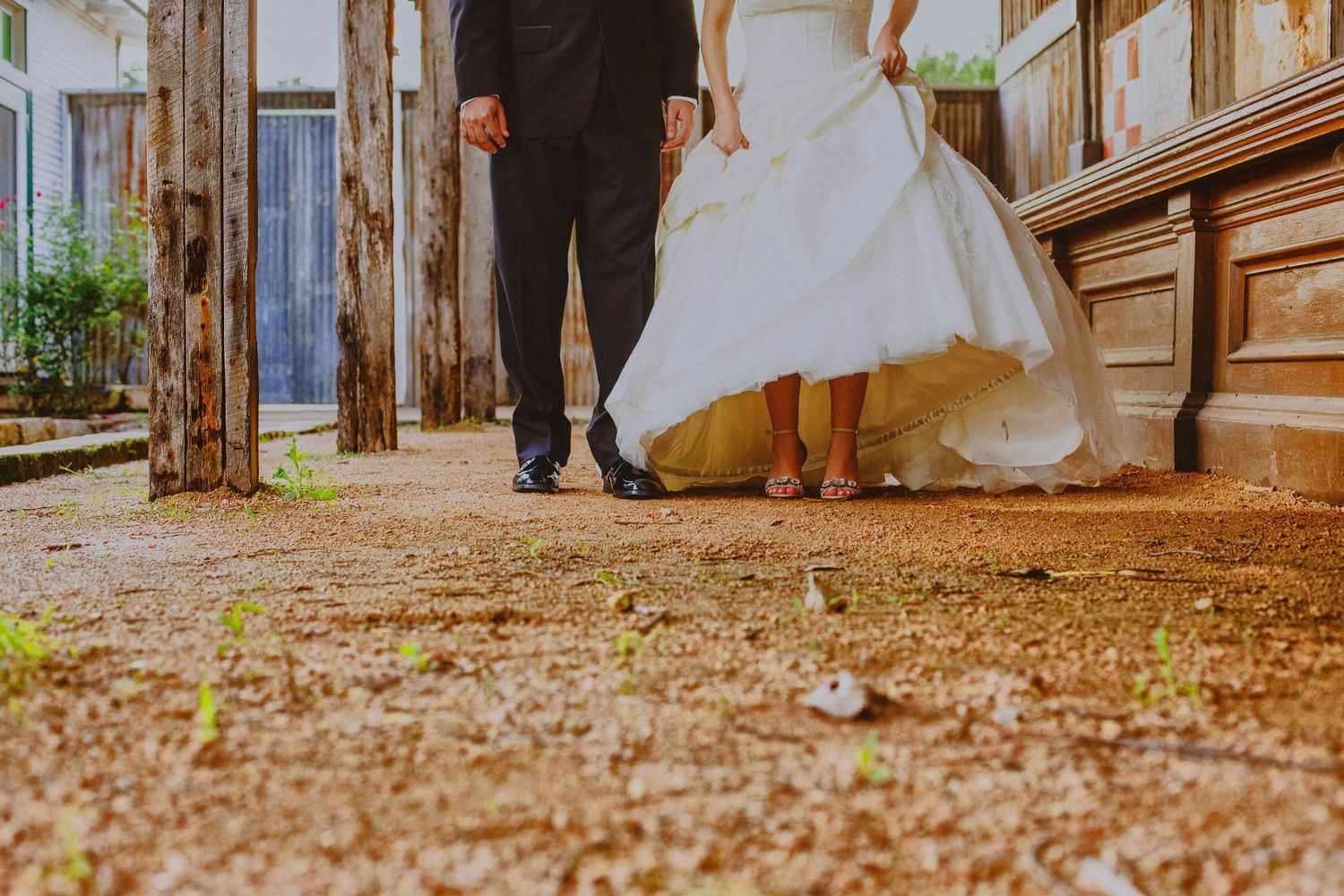 Bride in white gown and groom in suit walking hand in hand on a rustic path outside a historic building.