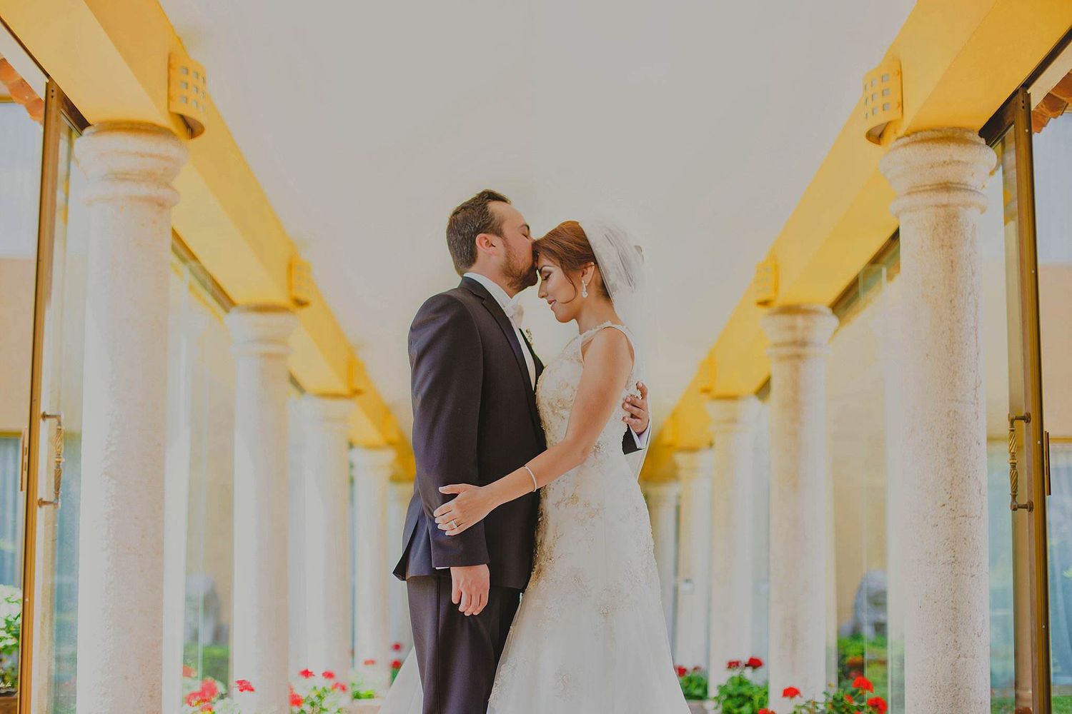 A newlywed couple shares a kiss under elegant white columns at their wedding venue.