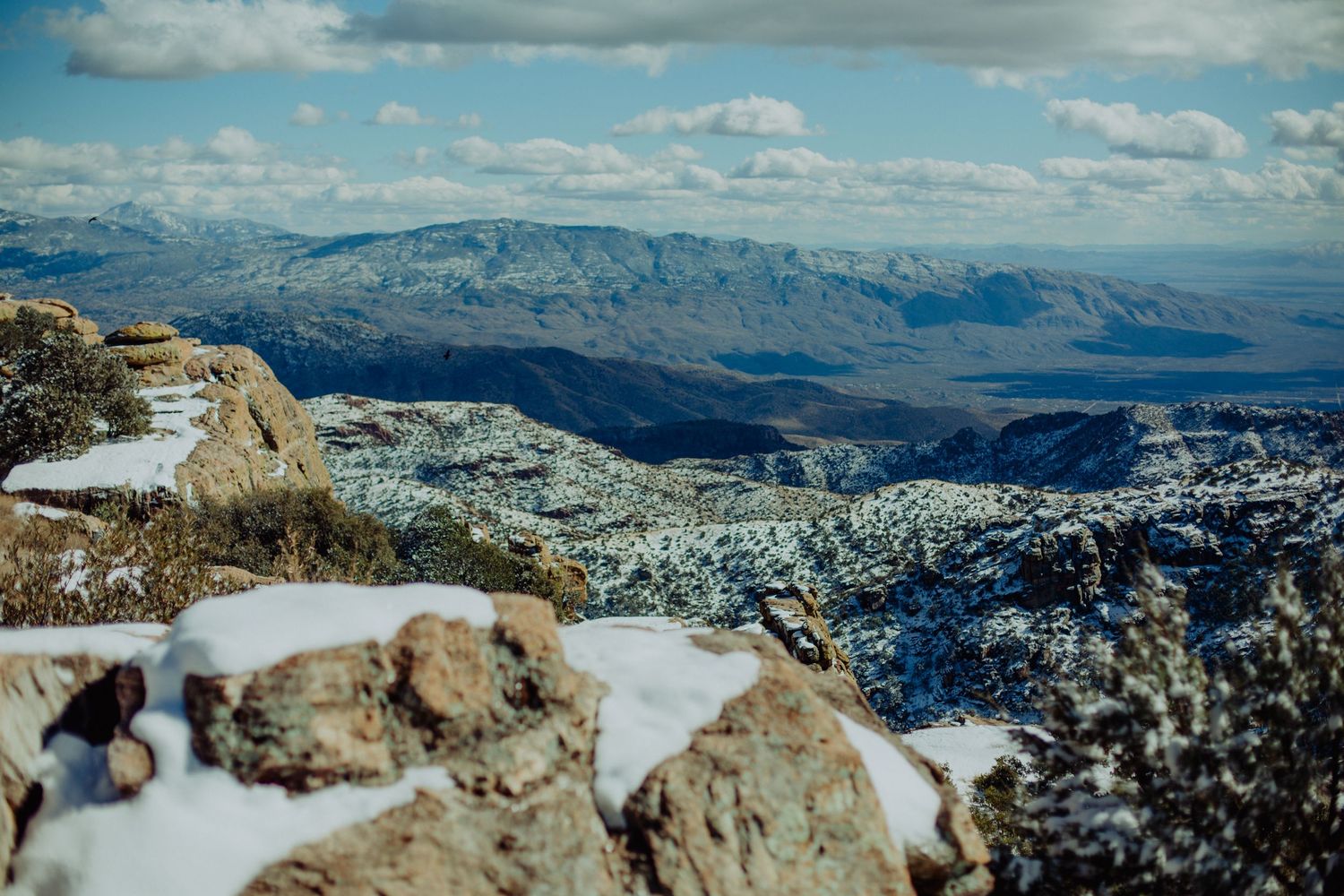 Snow-dusted rocky mountain summit with sweeping panoramic views of valleys and distant mountain ranges under a cloudy sky.