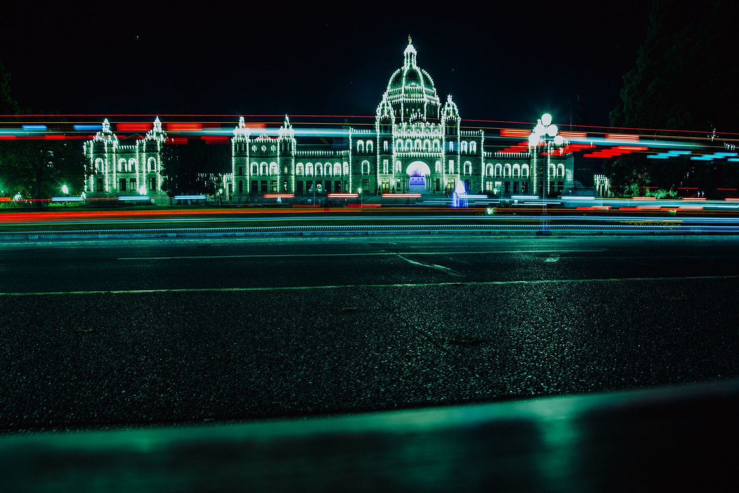 Night view of illuminated parliament building with light trails from passing cars on wet road.