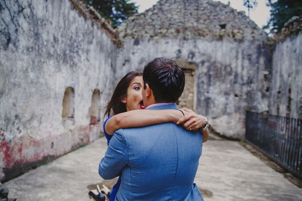 Couple embracing romantically in front of a rustic stone building, woman looking surprised and joyful.