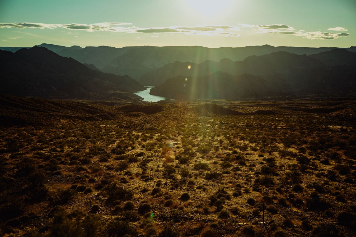 Aerial desert landscape at golden sunset with a winding river, rugged mountains, and sparse shrubs under a glowing sky.