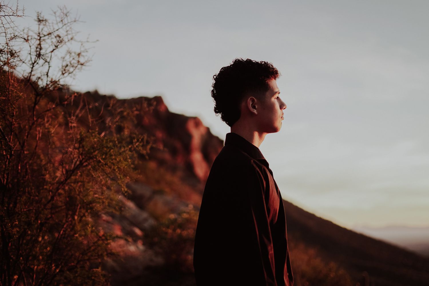 Young man in dark jacket stands in profile against rocky desert landscape at dusk with warm red tones.