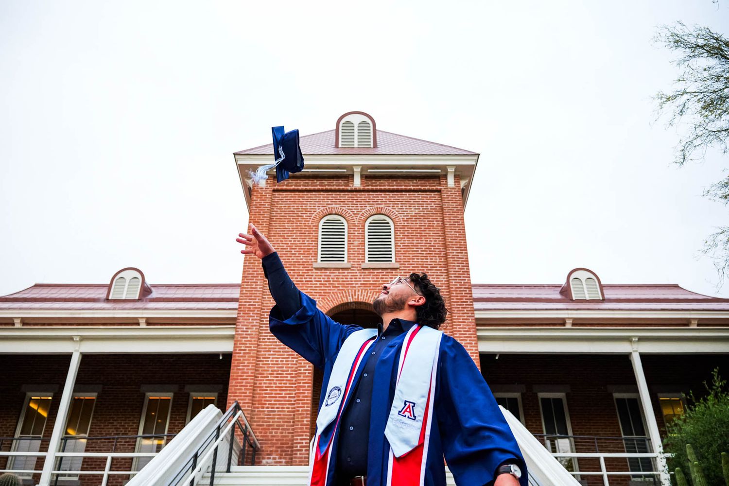 Graduate in cap and gown tosses mortarboard in front of historic brick university building.