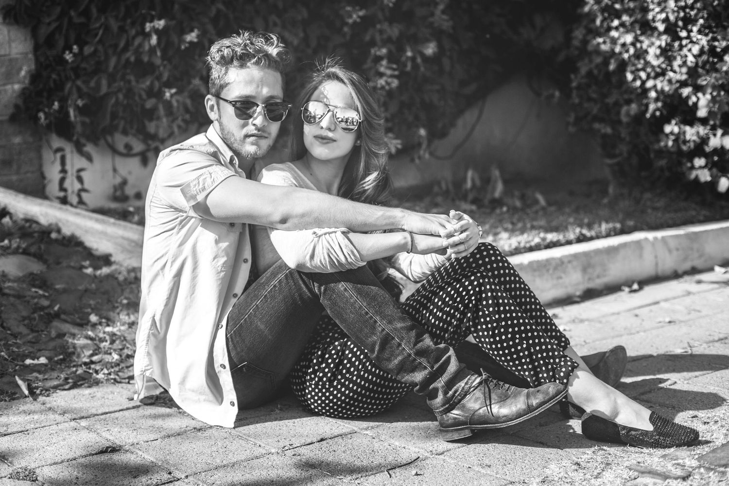 Black and white photo of a stylish couple sitting on the ground outdoors, both wearing sunglasses and casual attire.