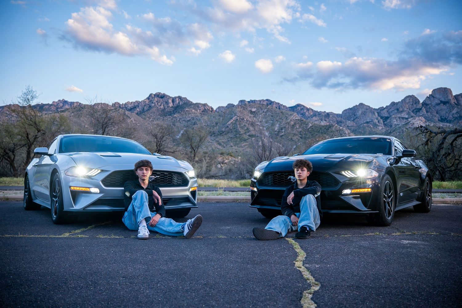 Two people sit in front of matching Ford Mustangs on a parking lot with rocky mountains at dusk.