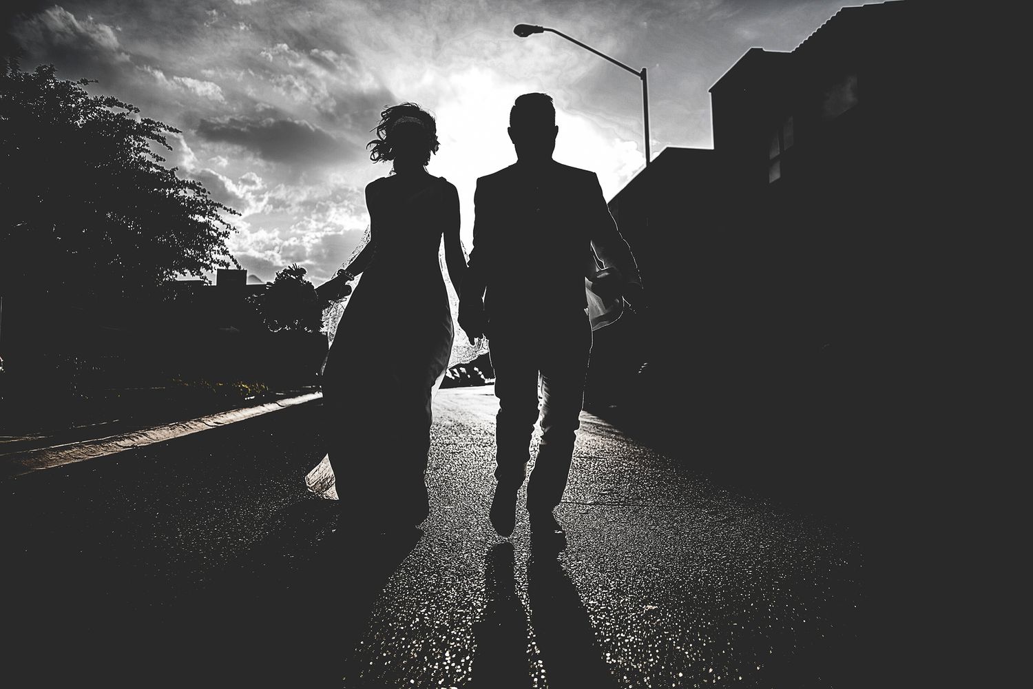 Silhouette of a bride and groom holding hands walking down a street at sunset on their wedding day.