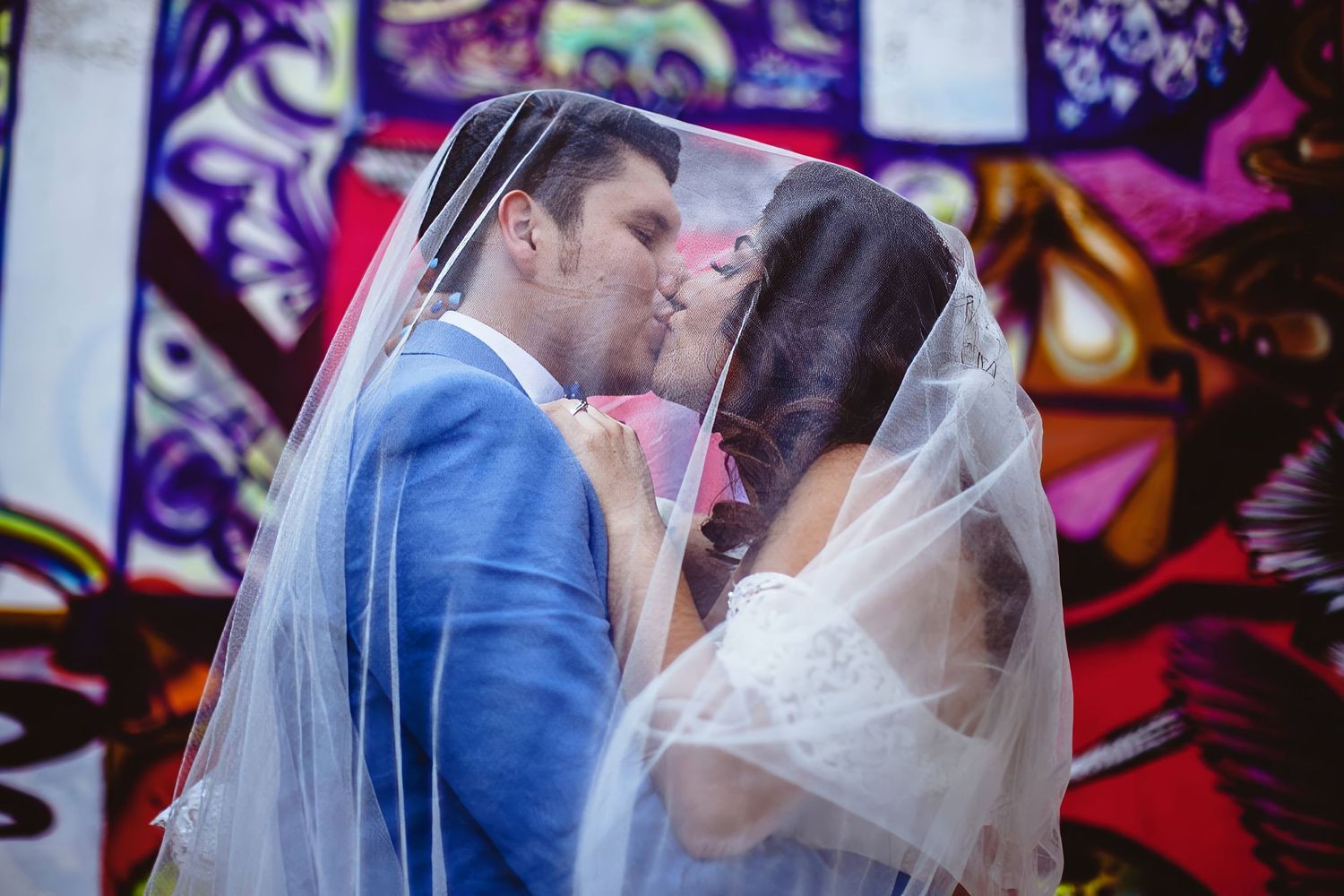 A newlywed couple shares a romantic kiss under a white veil in front of a colorful graffiti wall.