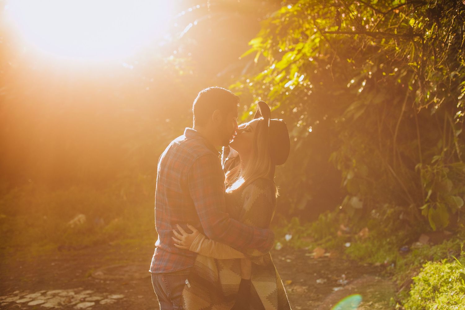A couple sharing a romantic kiss outdoors bathed in warm golden sunset light among green trees.