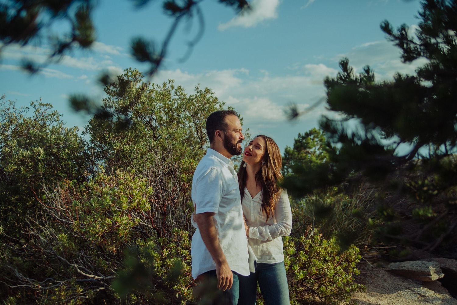 Couple standing among lush green trees and shrubs under a blue sky, sharing a romantic outdoor moment.