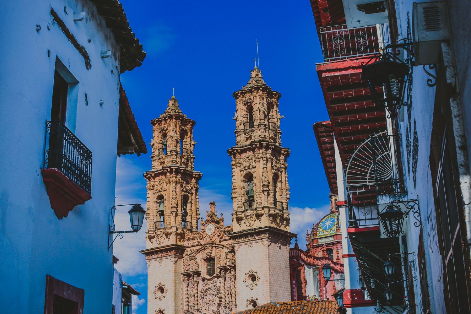 Ornate baroque church towers rise above colorful colonial buildings under a vivid blue sky in Taxco, Mexico.