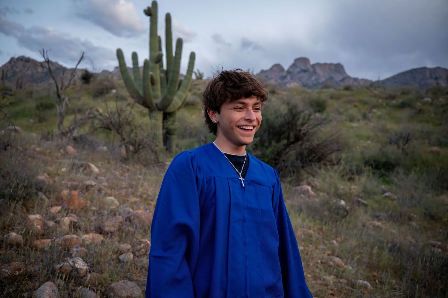 Young man in blue graduation gown smiling outdoors in Arizona desert with saguaro cactus and mountains behind him.