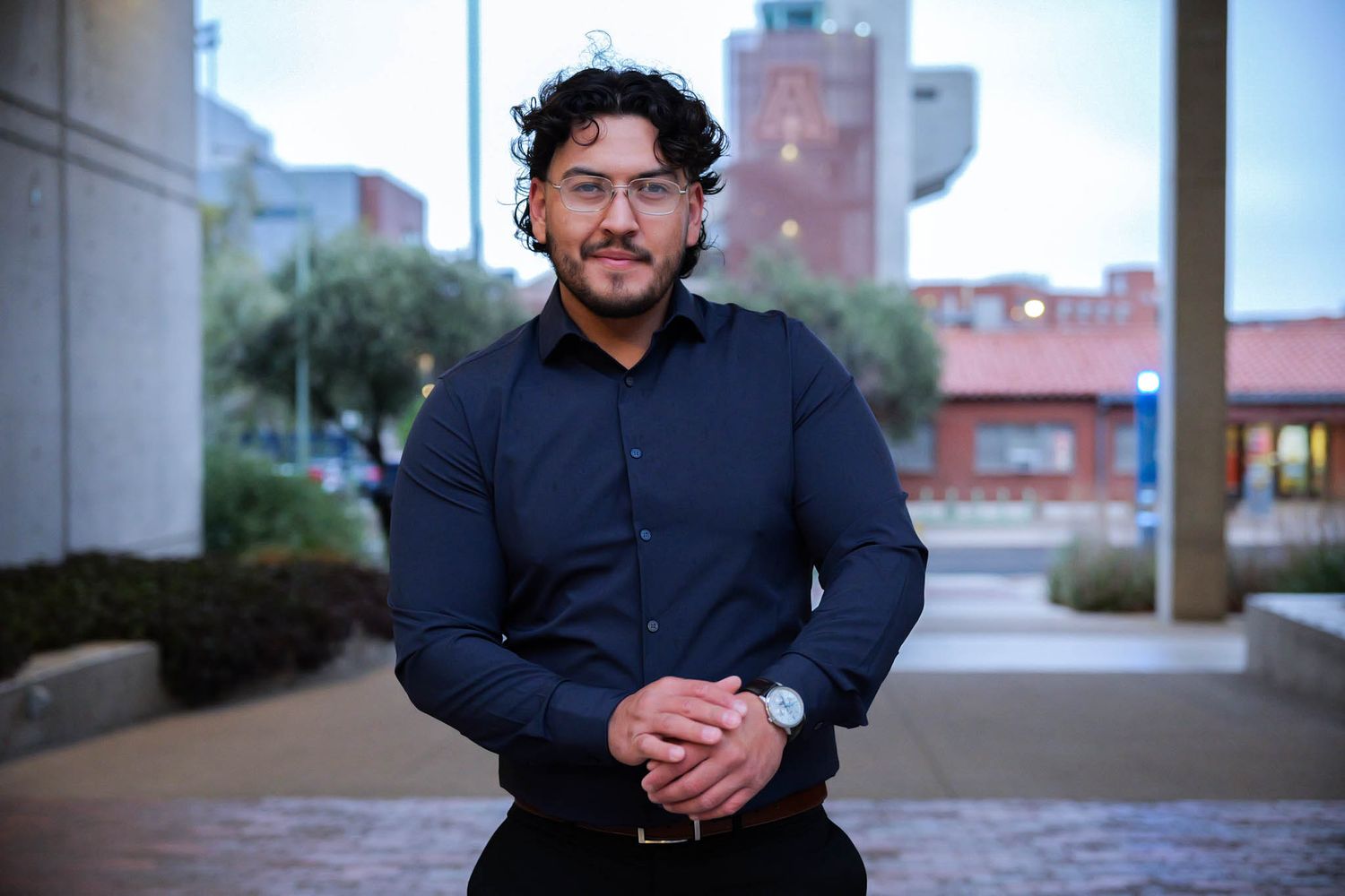 Professional young man in dark shirt poses outdoors near modern building in urban setting.
