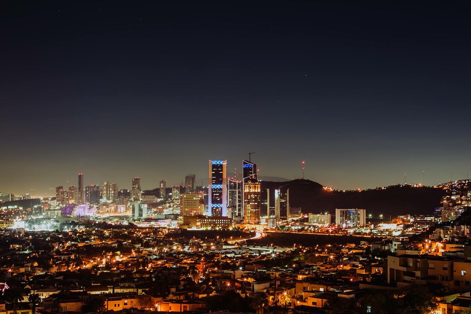 Aerial night view of a glowing city skyline with illuminated skyscrapers and urban lights under a dark blue sky.