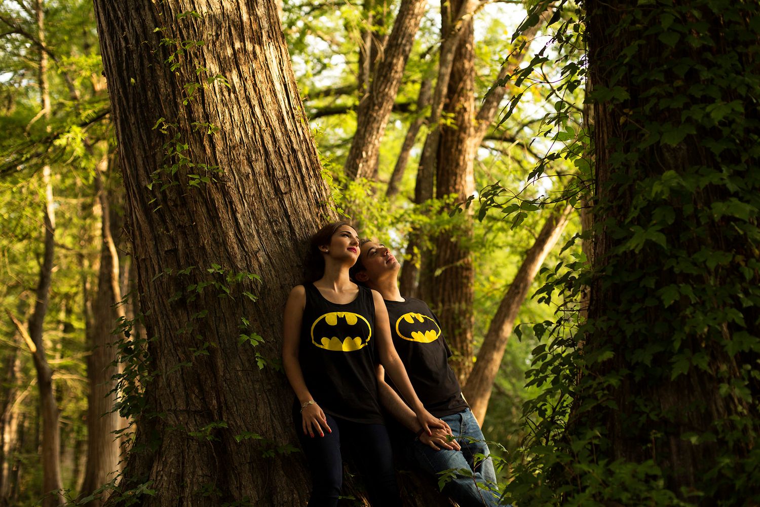 Two people in matching Batman shirts lean against a large tree in a lush green forest, gazing upward.