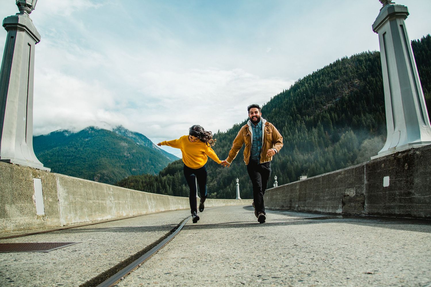 Couple holding hands running joyfully on a bridge with mountain and forest backdrop on a cloudy day.