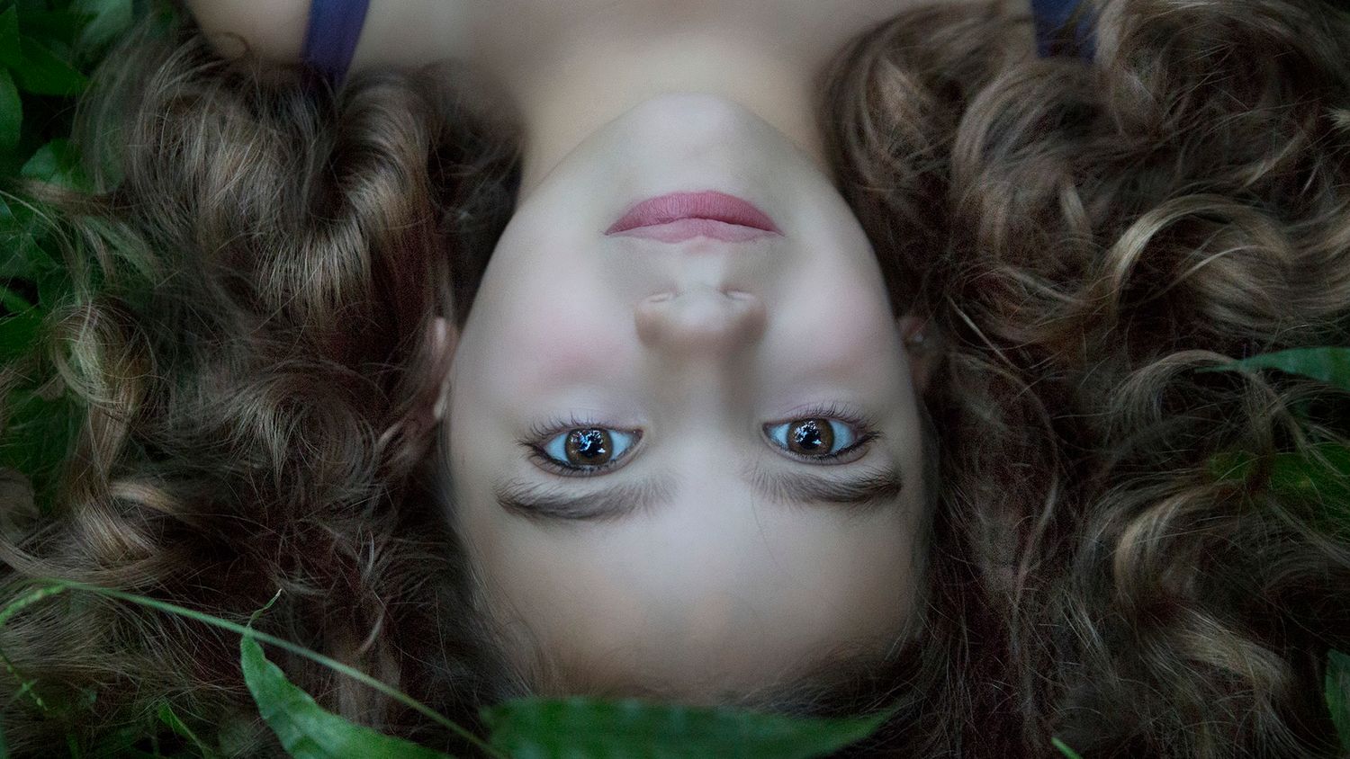 Young woman with curly brown hair lying on grass, looking up with blue eyes and natural makeup.