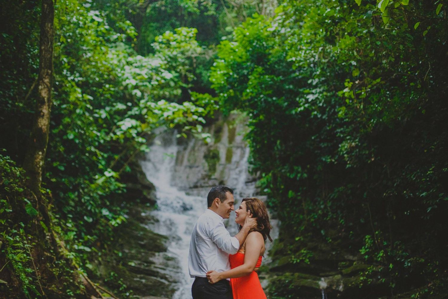 Couple embracing romantically in front of a lush tropical waterfall surrounded by green foliage.