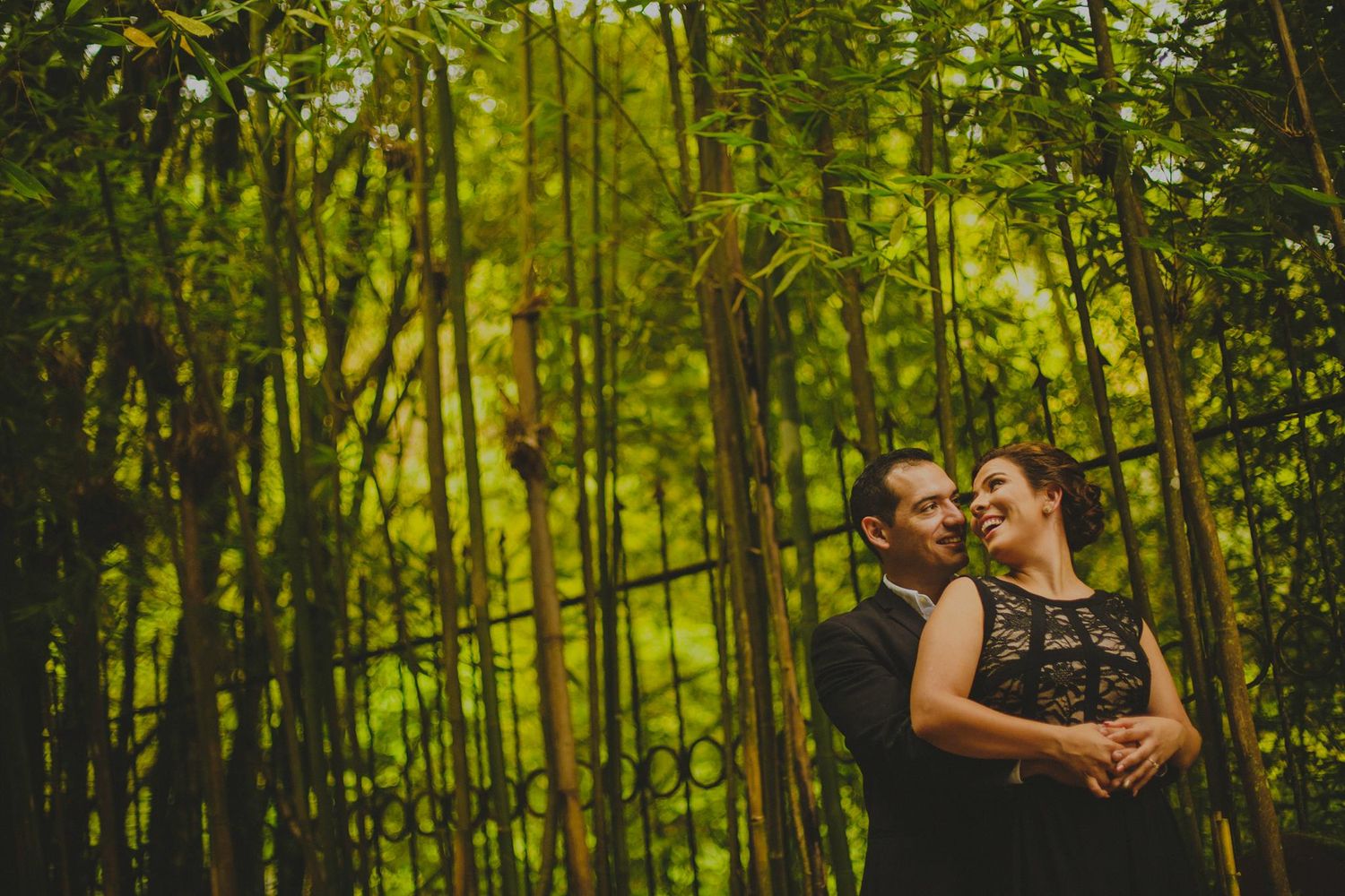 Couple in black formal attire embracing romantically among lush green bamboo trees in an outdoor garden setting.