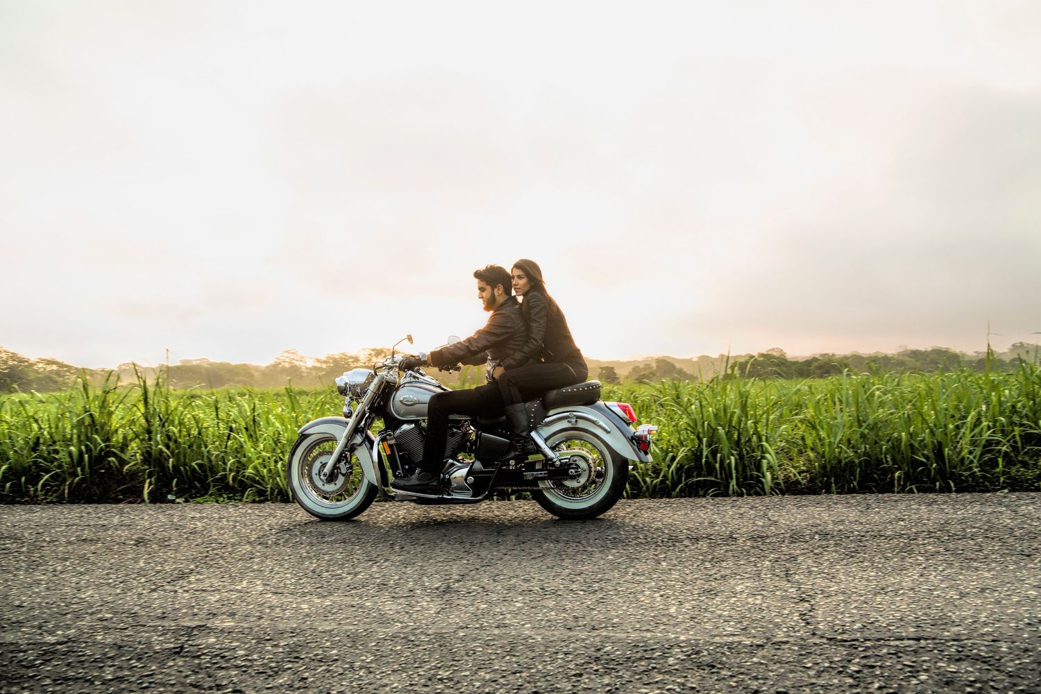 Two people riding a classic motorcycle together on a road beside lush green fields at golden hour.