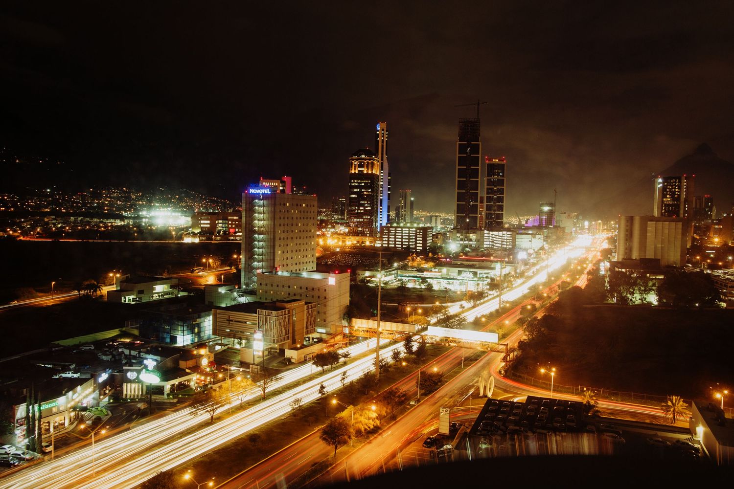 Aerial night view of a vibrant city skyline with illuminated skyscrapers and light trails on busy highways.