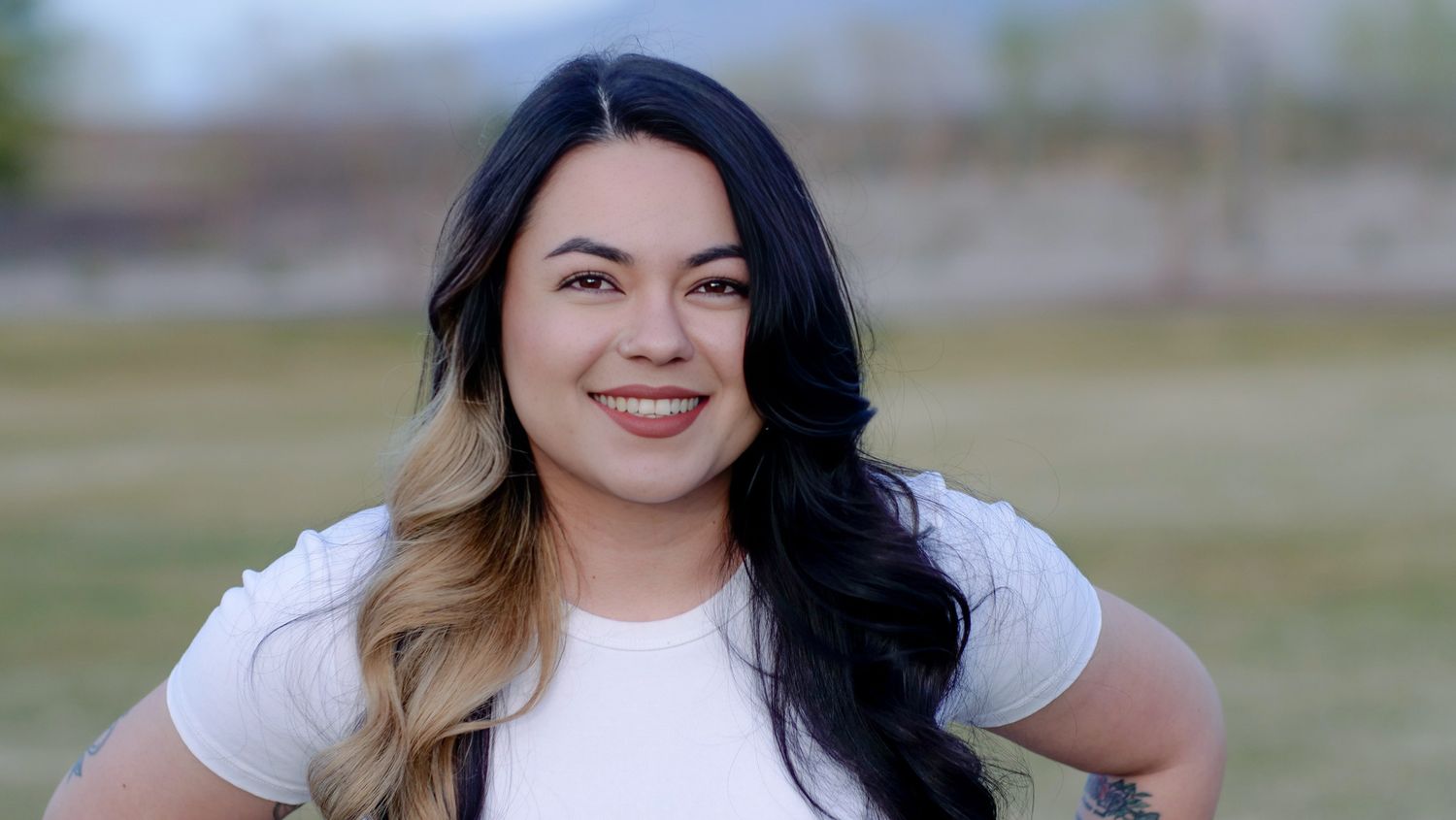 Smiling young woman with long black ombre hair wearing a white top, posing outdoors in a green field.