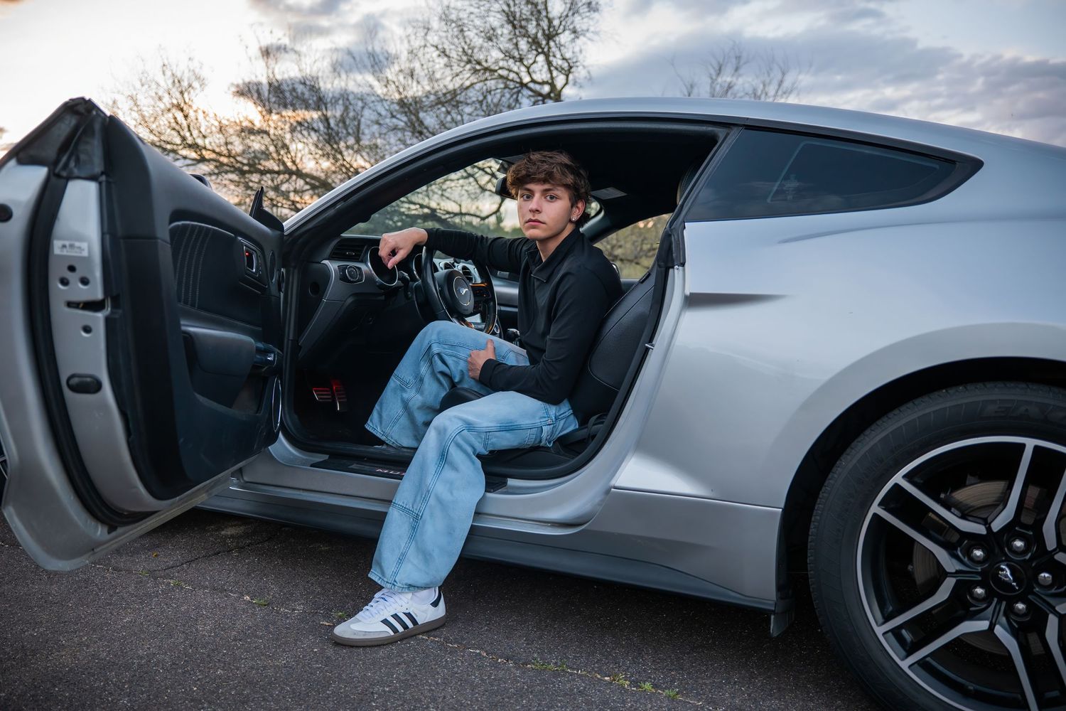 Young man in jeans and black jacket sitting in open door of silver Ford Mustang at dusk.
