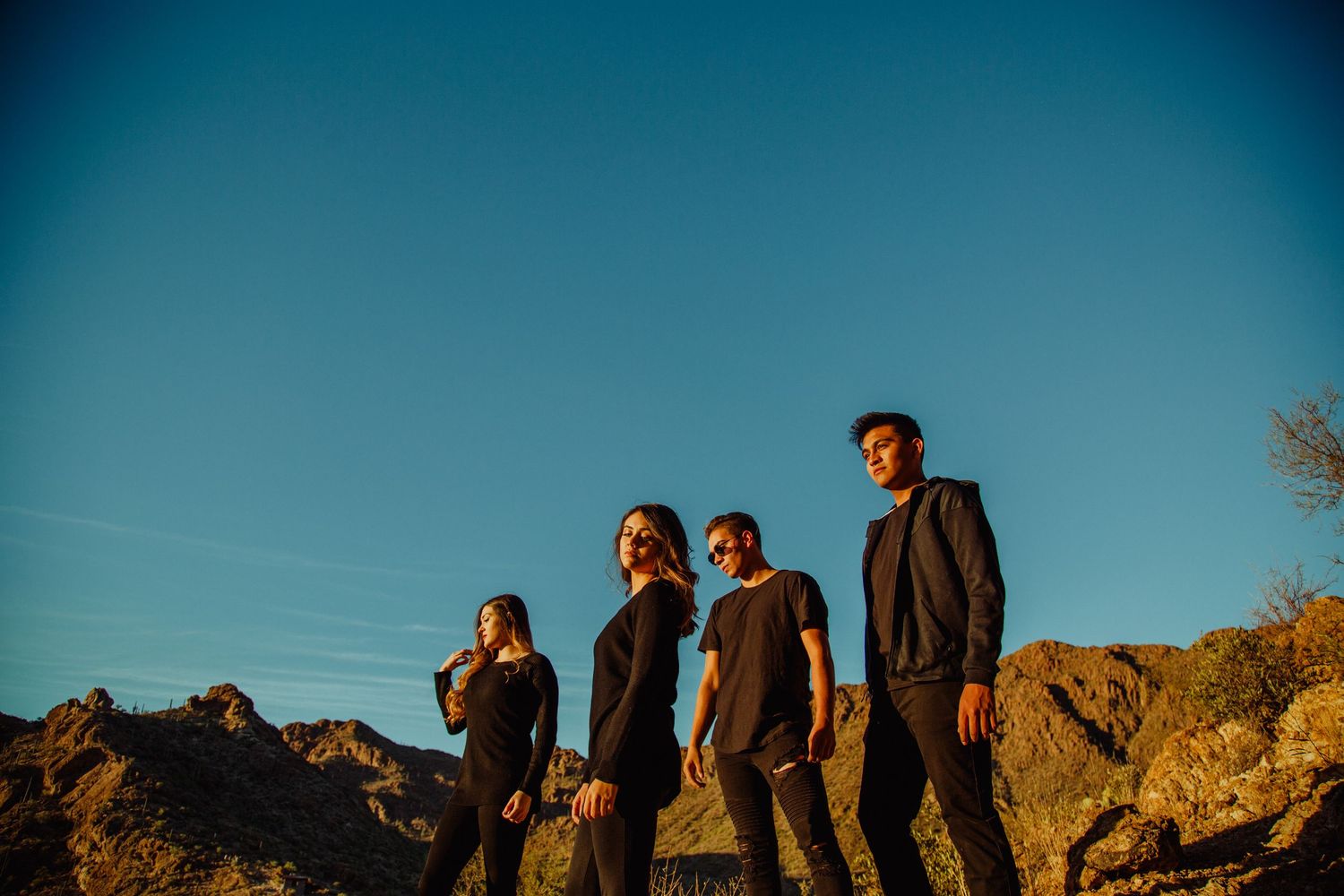 Four people in black clothing pose on rocky desert terrain under a vivid blue sky at golden hour.